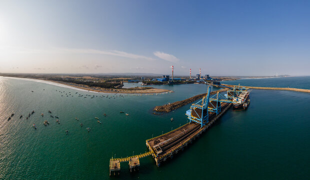 Aerial view of the coal terminal and power plant, featuring industrial cranes, a cargo ship, and small fishing boats in coastal waters in Cilacap, Central Java, Indonesia.