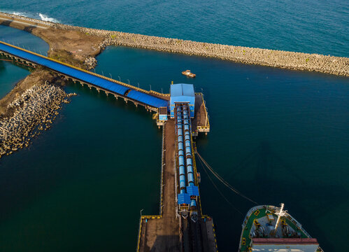 Aerial view of a coal terminal with a conveyor belt system extending into the sea, a cargo ship docked at the pier, and breakwaters in Cilacap, Central Java, Indonesia.