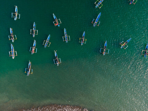 Aerial view of traditional outrigger fishing boats floating on the clear green water near the sandy shore in Cilacap, Central Java, Indonesia.