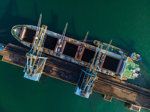 Aerial view of a large cargo ship docked at a port being loaded with coal by industrial cranes in the harbor of Cilacap, Central Java, Indonesia.