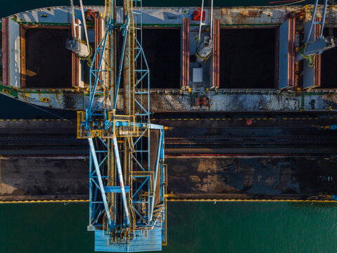 Aerial view of a cargo ship docked at a port with a large loading crane extending over open holds, industrial infrastructure, and harbor waters in Cilacap, Central Java, Indonesia.
