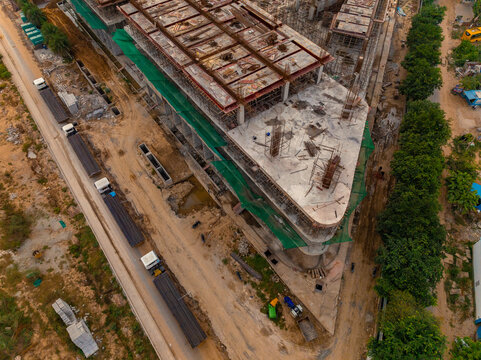 Aerial view of a large construction site with concrete frameworks, steel beams, green safety netting, and trucks parked on a dusty road in Gurugram, Haryana, India.