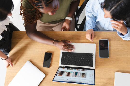 Three diverse women sit together at a wooden office desk, collaborating on a project. One person uses a laptop to search for ideas while their peers look on.