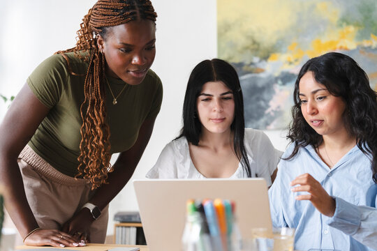 Three diverse female coworkers gather around a laptop in a bright office to collaborate on a project, discussing digital content together and sharing ideas.