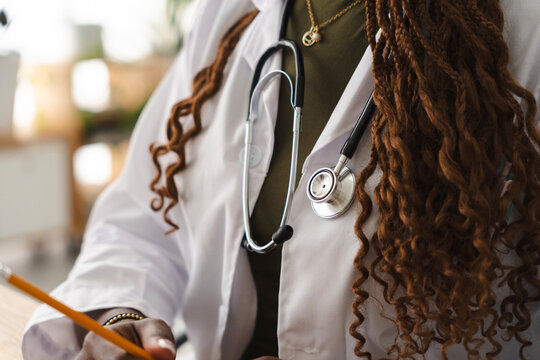 A female medical professional with long braided hair wearing a white coat and stethoscope sits at a desk while writing patient notes with a yellow pencil during a consultation.