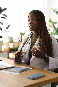 A Black female doctor wearing a white coat and stethoscope sits at her desk. She holds a pen and gestures while explaining medical results to a patient in her office.