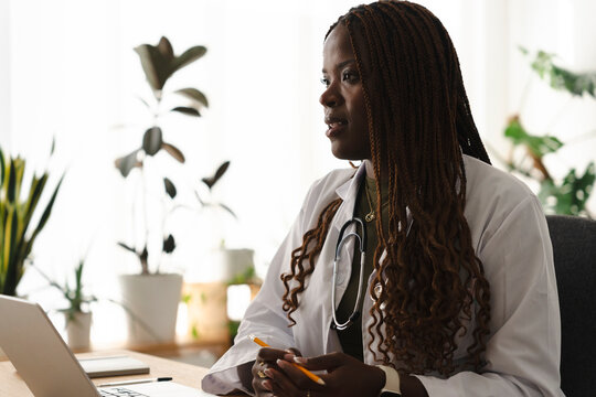 A Black female doctor wearing a white coat and stethoscope sits at a desk, holding a pencil while participating in a virtual consultation on a computer in a bright office.