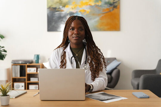 A Black female doctor wearing a white coat and stethoscope sits at her office desk. She works on a laptop while looking directly at the camera with a calm and confident expression.