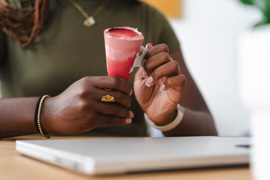 A Black woman sits at a wooden desk with a laptop computer and unwraps a red packaged ice cream cone. She carefully pulls the paper off the frozen treat during a work break.
