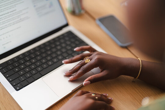 A Black woman works at a wooden desk, using the trackpad of her laptop. Her hands feature elegant rings and bracelets while she navigates a document on the screen.