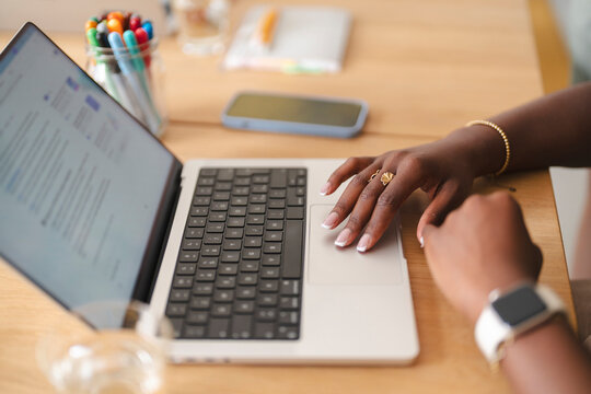 A young Black woman sits at a wooden desk in a modern office, actively working and using the trackpad of a laptop. Colorful pens and a mobile phone rest nearby.