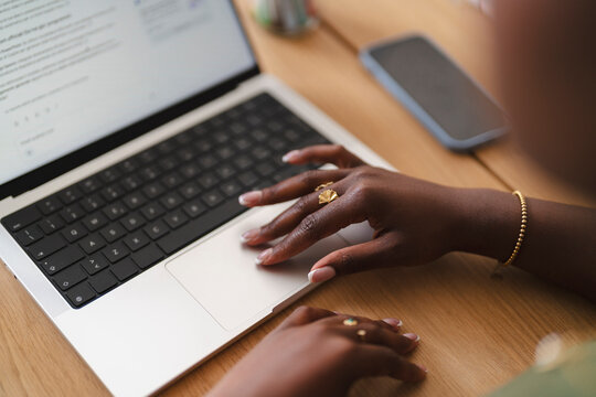 A Black woman sits at a wooden desk working on her laptop. Her hands rest on the keyboard and trackpad while she focuses on her daily remote work tasks.
