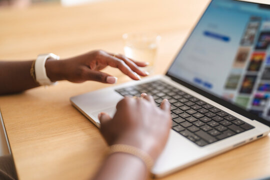 A Black woman typing on a laptop keyboard at a wooden desk. She works remotely on a modern computer while browsing a website in a bright workspace today.