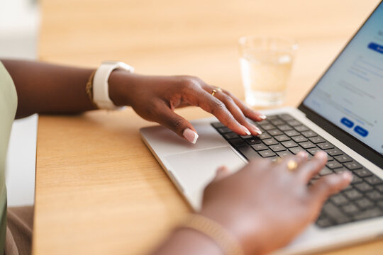 A black woman sits at a wooden desk and types on a modern laptop computer. Her hands are positioned over the keyboard while she works on a digital task.