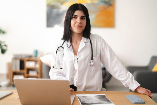 A young female doctor wearing a white coat and stethoscope stands confidently behind her office desk, looking at the camera with a gentle smile while working on a laptop.