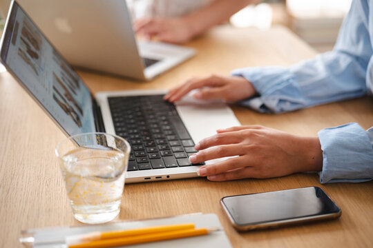 Two professionals work together at a bright wooden office desk. A young woman types on a laptop computer while reviewing online content, with a glass of water and a phone nearby.