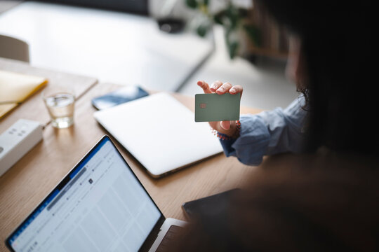 A person sits at a wooden desk with a laptop and holds up a bank card. They are making an online purchase or reviewing financial details in a bright modern home office.