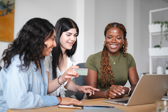 Three diverse young women sit together at a wooden desk and smile while shopping online. One woman holds a credit card up to pay for a purchase on an open laptop computer.