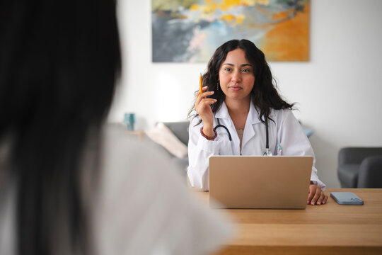 A female doctor wearing a white coat and stethoscope sits at a desk with a laptop, listening attentively to a patient during a medical consultation in a bright office.