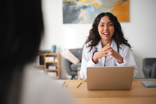 A smiling female doctor in a white coat sits at her office desk with a laptop, engaging warmly in a consultation with a patient seated across from her in the medical clinic.