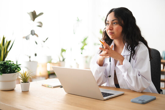 A young female doctor in a white coat sits at a wooden desk with a laptop, thoughtfully listening during a telehealth consultation in a bright plant-filled office.