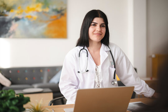 A young female doctor wearing a white lab coat and stethoscope smiles warmly while sitting at her desk in a modern office, ready to consult with patients.