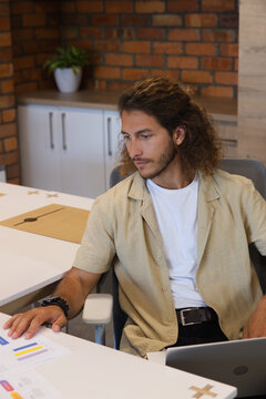 Man sitting at office desk wearing beige short-sleeve shirt, reaching printed charts and laptop
