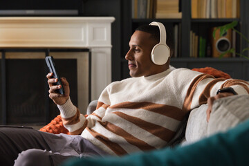 Fototapeta premium African American man reclining on gray couch, wearing white headphones, holding smartphone
