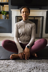 Naklejka premium African American woman meditating on black yoga mat in living room, wearing leggings and smartwatch