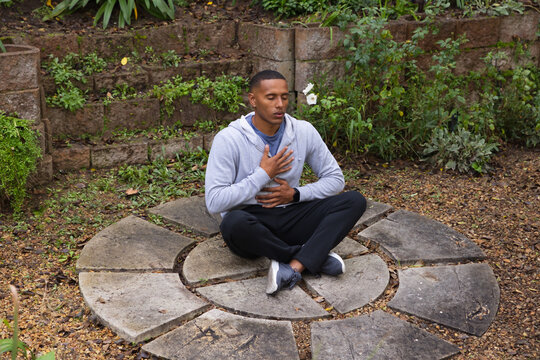 African American man sitting cross-legged meditating on stone ring in garden with hoodie smartwatch