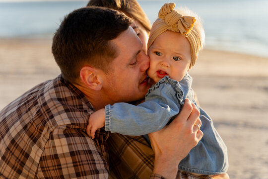 A loving father kisses his baby daughter on the cheek during a sunny day at the sandy beach
