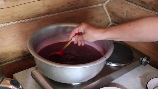 Making redcurrant jelly. A woman's hand slowly stirs the mixture clockwise with a wooden spoon in a metal basin on an electric hot plate in a country house. Homemade berry preserves &ndash; horizontal video