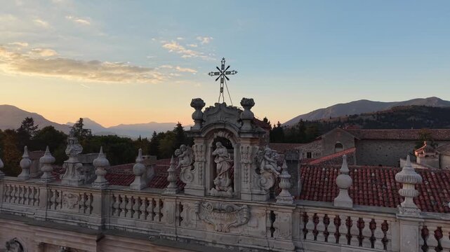 Baroque statue and iron cross on the roofline of the Certosa di Padula against a golden sunset sky, Campania, southern Italy