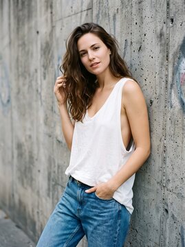 Casual Brunette Woman Leaning Against Concrete Wall Wearing White Tank Top and Blue Jeans