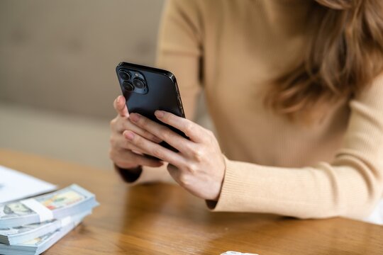 Woman using smartphone while sitting at wooden table with stack of money
