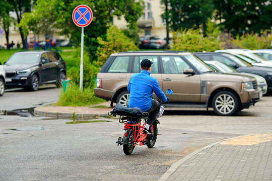 Pizza delivery courier in blue jacket riding red electric cargo bike along street near parked cars in residential district, delivery service. Food delivery service, man on e-bike delivers food orders