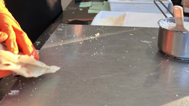 Hands in orange gloves are preparing borlengo, a traditional Italian street food pancake from the Emilia Romagna in Italy, filling it with a savory mixture and Parmigiano cheese on a metallic surface