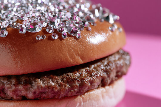 Close-up gourmet burger with a sparkling crystal-covered bun and juicy beef patty against a vibrant pink background.
