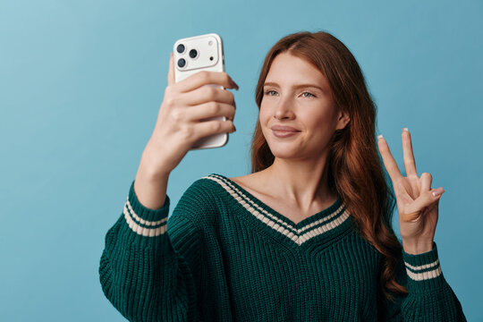 Smiling woman in a green sweater taking a selfie with a smartphone against a blue studio background. Casual portrait conveying wellness, confidence, and everyday style. Soft lighting creates a warm