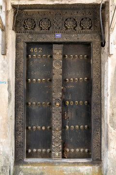 Ornate carved wooden door number 462 with brass studs and a floral carved frame and lintel set in a weathered stone wall with a blue house number plate above, Stone Town, Tanzania