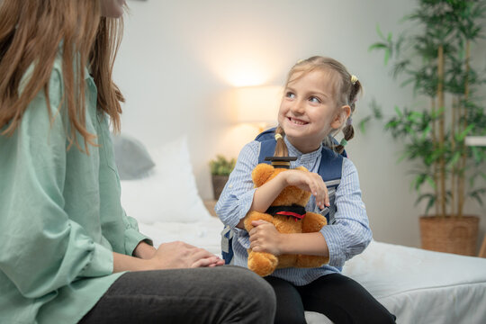 Happy little girl holding teddy bear and looking at her mother with a smile. Cheerful elementary schoolgirl wearing backpack holding graduation teddy bear and smiling at supportive mother in bedroom.