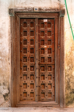 Old wooden door with square panels and iron studs set in a moulded frame on a peeling plaster wall with a green cable running down the wall, Stone Town, Tanzania