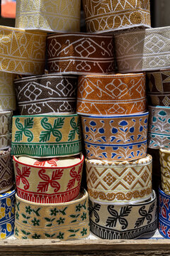 Stack of colorful embroidered kofia caps with floral and geometric patterns displayed on a rustic wooden shelf in bright midday sunlight in a market, Stone Town, Tanzania