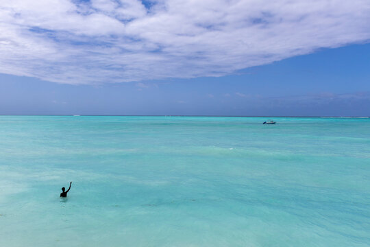 Person standing waist deep in calm turquoise water raising an arm in greeting with a small speedboat anchored beyond and soft clouds over the horizon, Nungwi, Tanzania