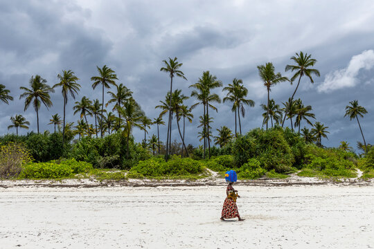 Woman in a colorful long dress carrying a blue bundle on her head as she walks along a white sand beach beneath coconut palms and stormy clouds, Matemwe, Tanzania