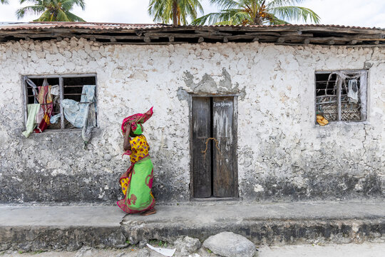 Woman in a bright green and yellow kanga carrying a bundle on her head walking past a whitewashed coral stone house with wooden doors and barred windows, Matemwe, Tanzania