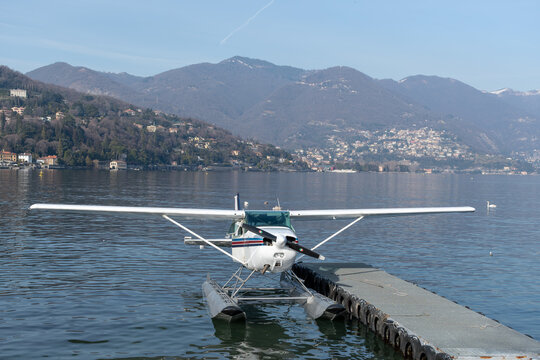Floatplane moored at a dock on Lake Como, framed by calm water, hillside towns and mountains beneath a clear blue sky in northern Italy.