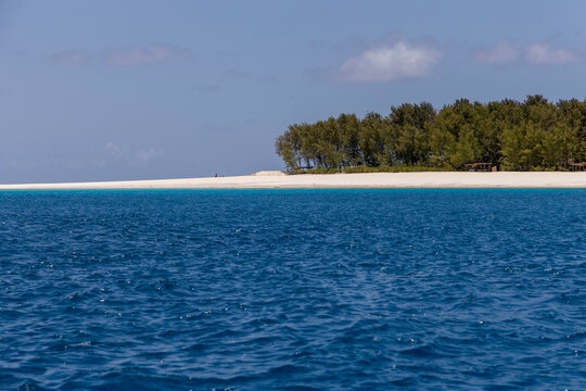 Small tropical island with green trees and a white sand beach seen across deep blue sea under a hazy blue sky with thatched shelters on the shore, Matemwe, Tanzania