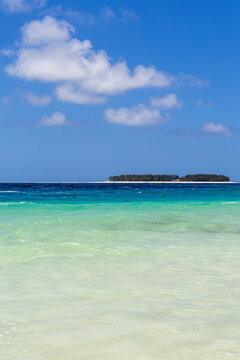 Vertical view of a small low island with green trees on the horizon beyond turquoise and emerald shallow sea water under a bright blue sky with soft white clouds, Matemwe, Tanzania