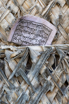 Old worn booklet with Arabic script tucked into a weathered woven palm leaf wall with frayed gray and beige strips and a knotted white cloth in a close view, Matemwe, Tanzania